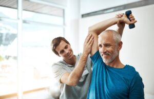 Mature man lifting dumbbell with guidance from physiotherapist, focusing on strength, mobility, and rehabilitation in a clinical setting.