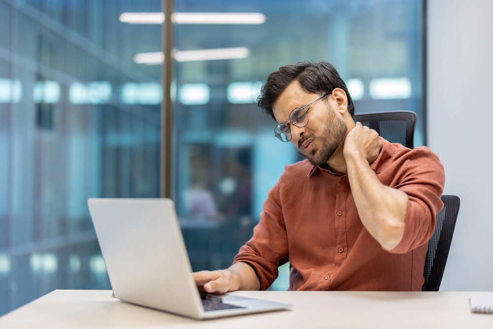Man experiencing neck pain while using a laptop in an office, illustrating orthopedic neck disorders and the impact of poor ergonomics during prolonged computer work.