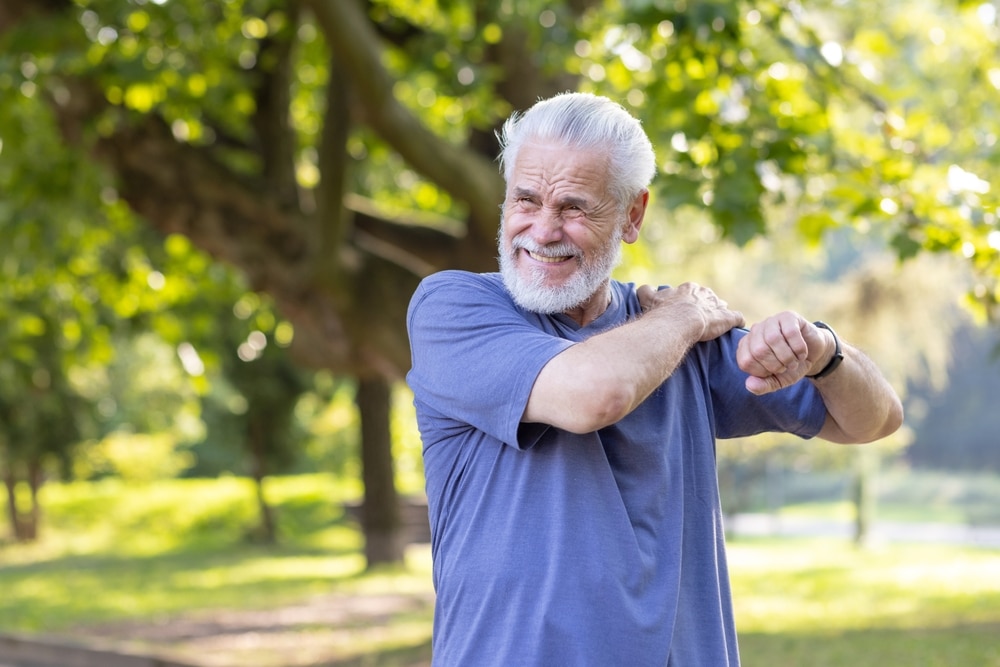 Senior man outdoors massaging shoulder in pain after ankle sprain while walking and exercising in park.