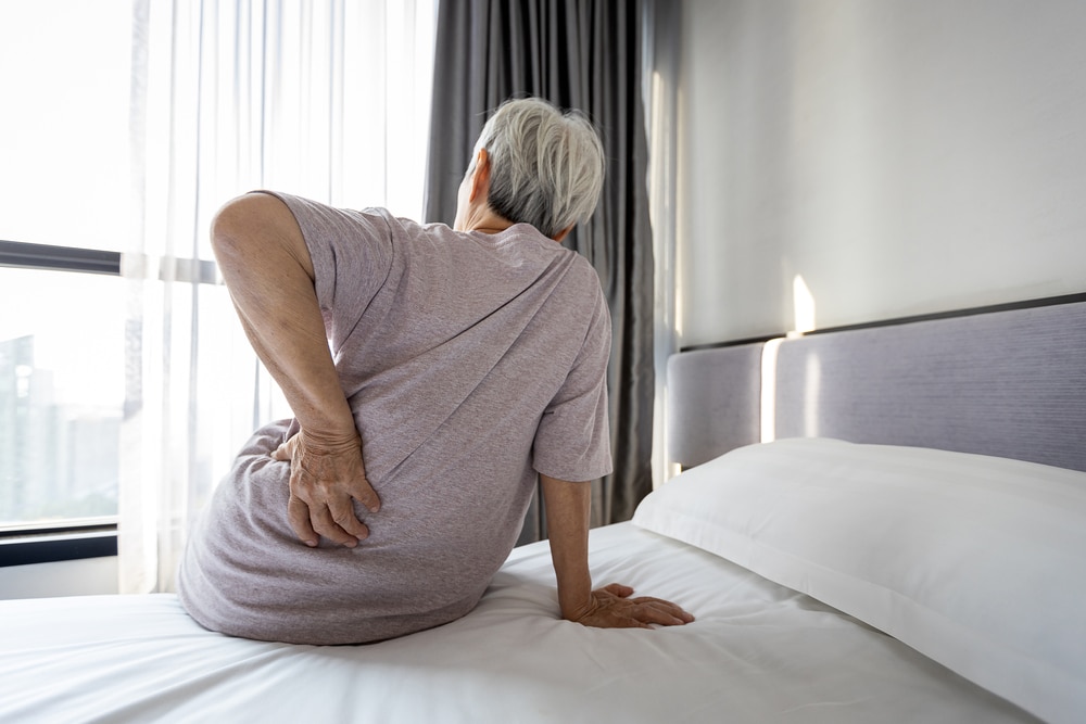 Senior woman sitting on bed massaging lower back due to hip pain from poor posture or uncomfortable mattress.