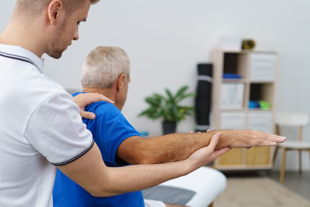 Physiotherapist treating man’s elbow injury by flexing the shoulder during rehabilitation therapy session in clinic.