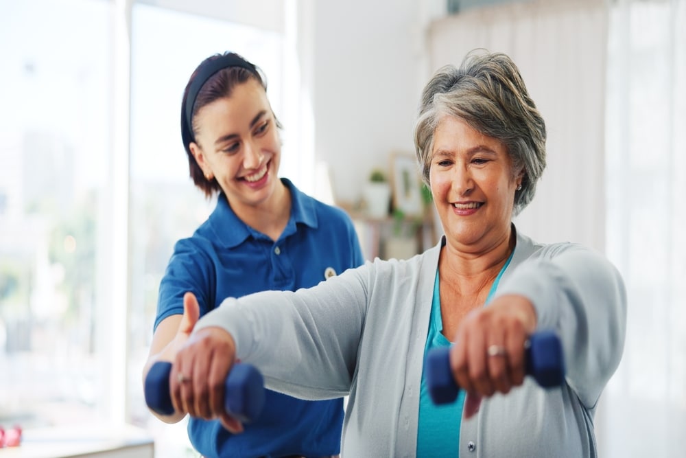 Smiling senior woman exercising with dumbbells assisted by therapist, concept of physiotherapy, rehabilitation, and mobility recovery